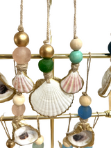 Hanging scallop shell ornaments with natural and painted beads in gold, green, turquoise, and blue, each edged with gold paint, displayed on a gold metal frame.