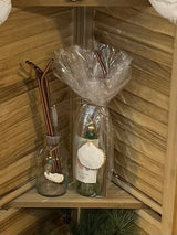 Two glass bottles with metallic straws, one wrapped in clear plastic and adorned with a hanging white scallop shell ornament, displayed on a wooden corner shelf.