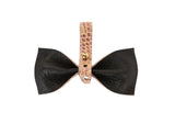 Bow-shaped object with black textured leather petals and a central strap in pink embossed croc pattern secured by a small gold button.