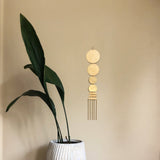 Wall hanging with five connected gold-toned circular metal discs above five hanging metal rods, displayed on a beige wall beside a tall green-leafed plant in a white textured pot.