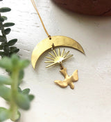 Gold-toned crescent moon pendant with a sunburst and dove-shaped charm hanging below, photographed on a white surface near green foliage.