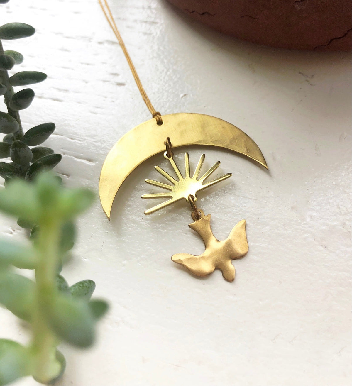 Gold-toned crescent moon pendant with a sunburst and dove-shaped charm hanging below, photographed on a white surface near green foliage.