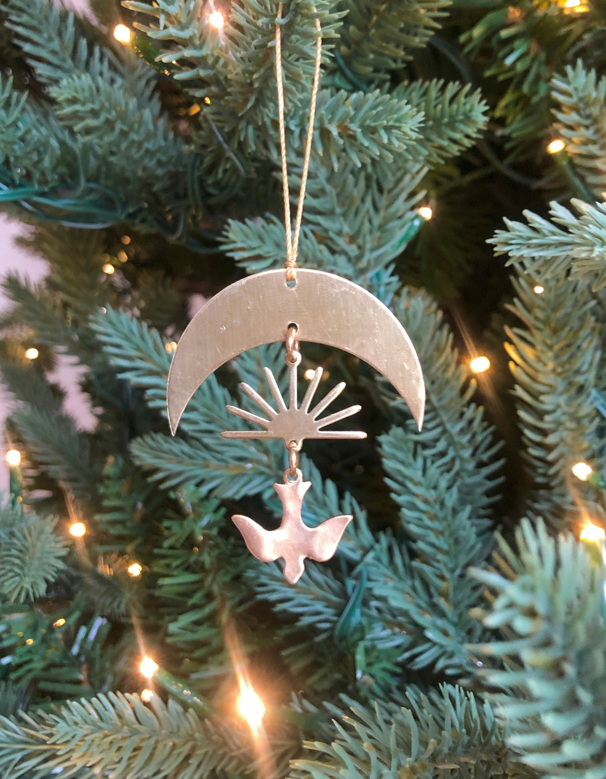 Metal ornament featuring a crescent moon shape with a sunburst and a dove hanging beneath, suspended on a Christmas tree with green needles and warm lights.