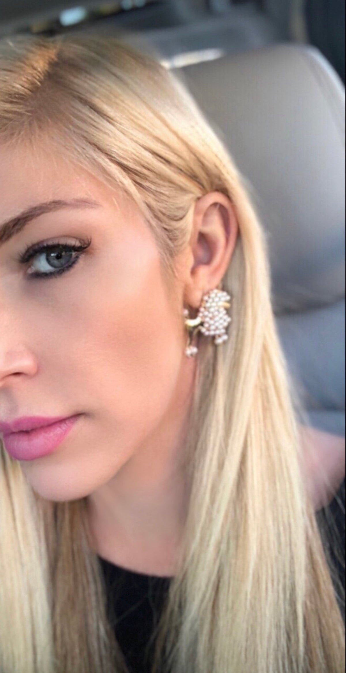 Close-up of a woman's left ear wearing a pearl cluster earring shaped like a poodle, with light blonde hair and subtle pink lipstick visible.