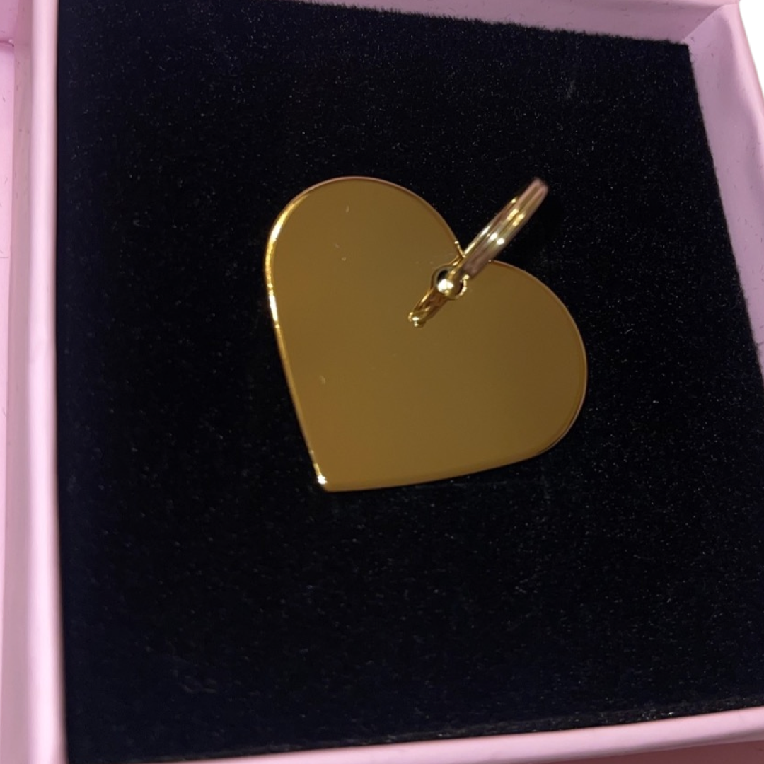 Gold heart-shaped charm with a small attached ring, placed in a black velvety square box with a pale pink border.
