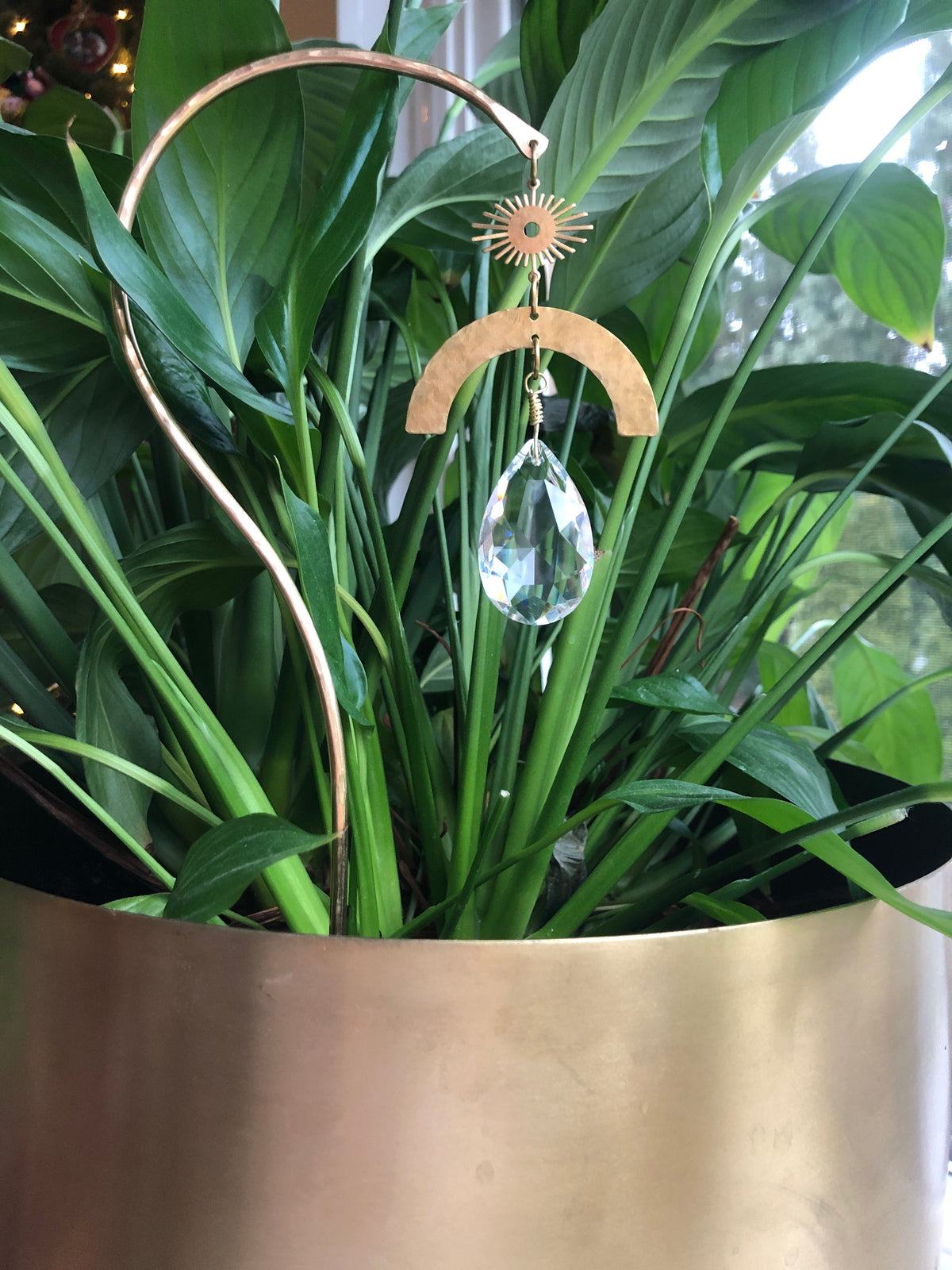 Gold-toned metal suncatcher with a sun shape, crescent, and teardrop crystal hanging amid vibrant green plant leaves in a gold planter.