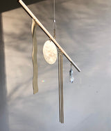Gold-toned metal suncatcher with a wavy strip, a round disk, three thin rods, and a faceted crystal pendant suspended from a slender horizontal bar against a muted background.