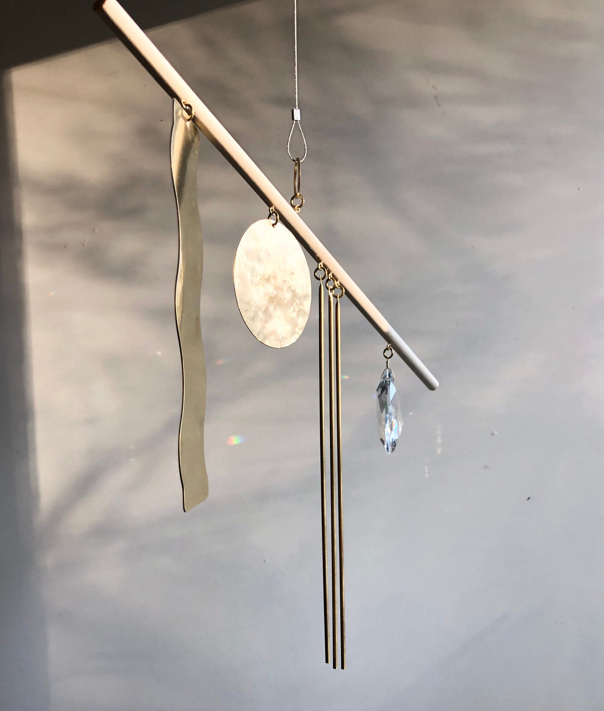 Gold-toned metal suncatcher with a wavy strip, a round disk, three thin rods, and a faceted crystal pendant suspended from a slender horizontal bar against a muted background.