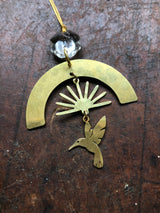 Brass ornament featuring a geometric arch with radiating rays above a stylized hummingbird, hanging from a vintage faceted glass crystal bead on a golden thread.