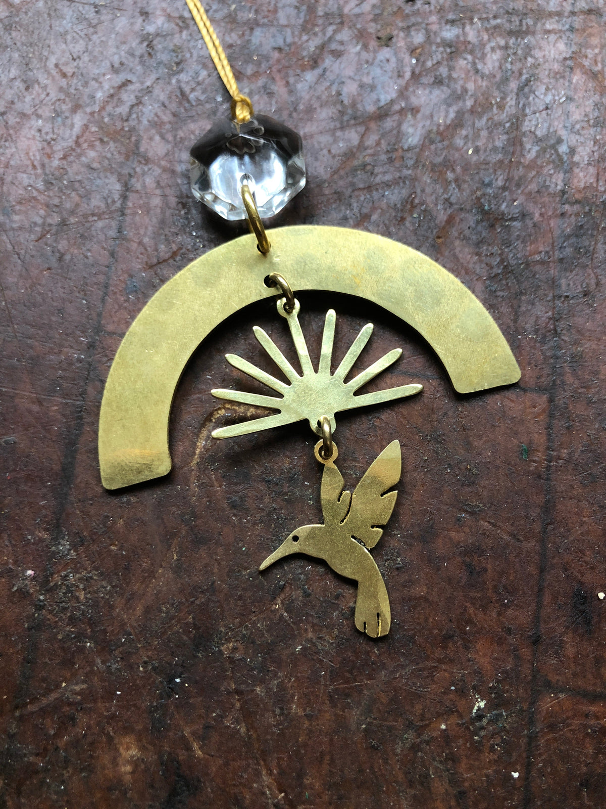 Brass ornament featuring a geometric arch with radiating rays above a stylized hummingbird, hanging from a vintage faceted glass crystal bead on a golden thread.