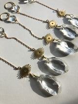 Three delicate sun catcher chains each with a gold sunburst charm and a large faceted clear vintage glass crystal pendant casting shadows on a white surface.
