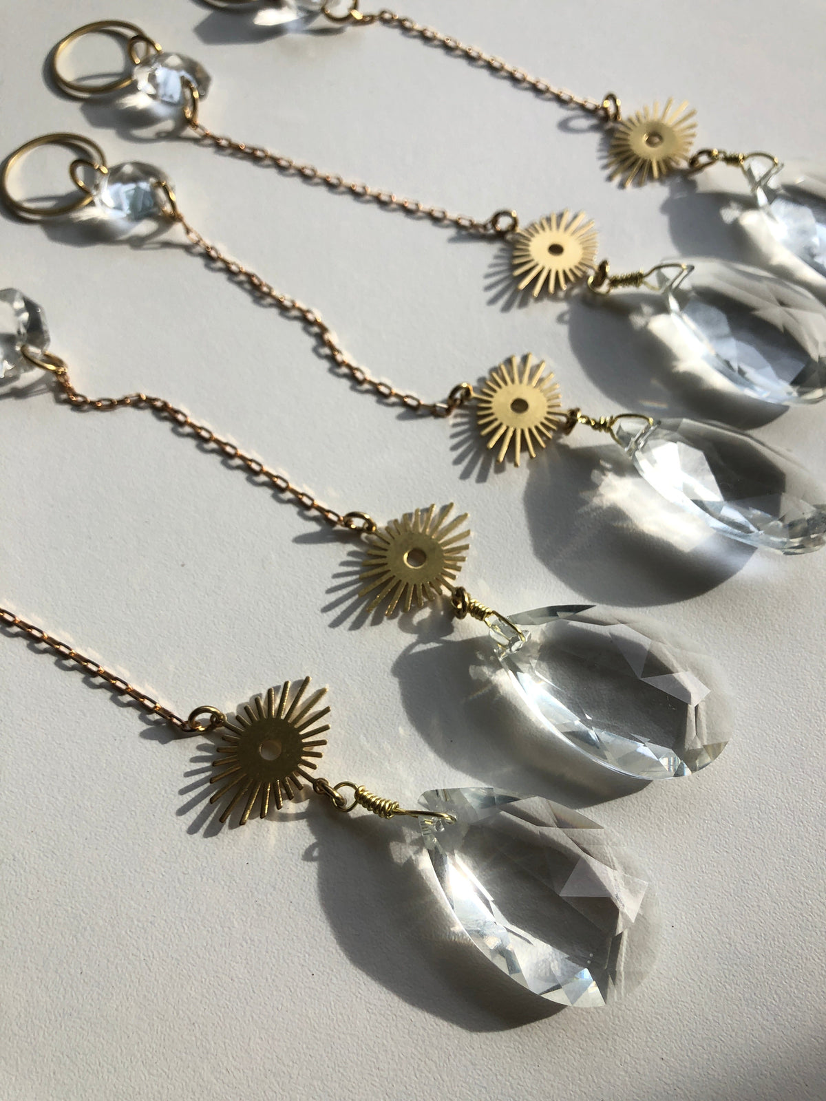 Three delicate sun catcher chains each with a gold sunburst charm and a large faceted clear vintage glass crystal pendant casting shadows on a white surface.