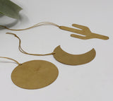 Three flat brass ornaments shaped as a cactus, crescent moon, and circle, each hanging by a thin twine loop on a white background.