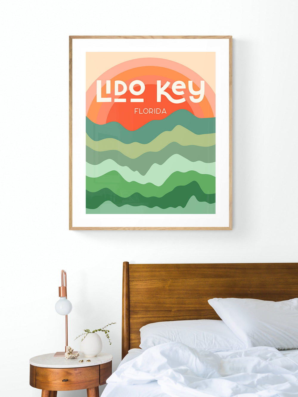 Modern art print with stylized layered waves in green tones beneath an orange sunset, featuring bold white text reading 'Lido Key Florida,' hung above a wooden bed.