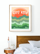 Modern art print with stylized layered waves in green tones beneath an orange sunset, featuring bold white text reading 'Lido Key Florida,' hung above a wooden bed.