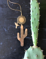 Gold-toned metal Desert Dream Ornament with a stylized sun, hummingbird, and cactus shapes on a thin gold string next to a green cactus pad with white areoles.