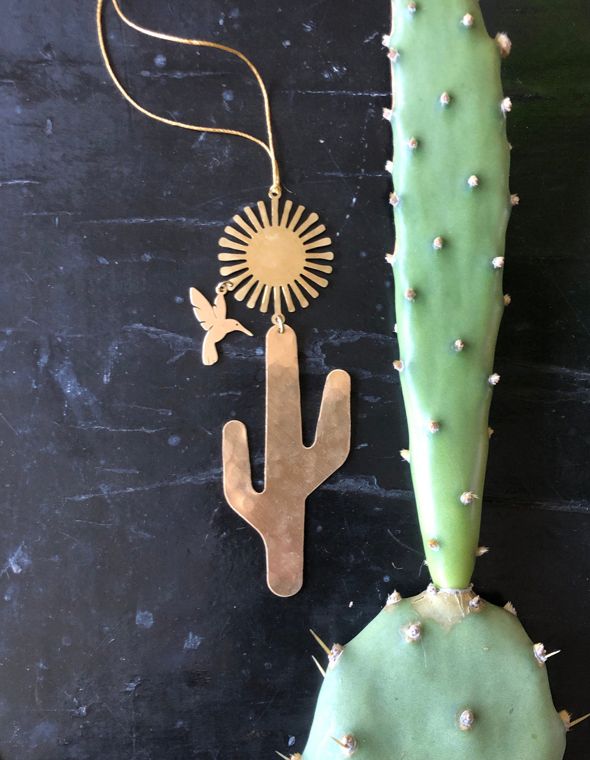 Gold-toned metal Desert Dream Ornament with a stylized sun, hummingbird, and cactus shapes on a thin gold string next to a green cactus pad with white areoles.