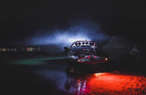 Black motorboat at night with red underwater LED lights shining in water and bright white lights mounted on the roof emitting foggy beams.