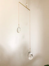 Minimalist suncatcher featuring two faceted crystal drops suspended from thin golden chains attached to a horizontal brass bar against a plain light wall.