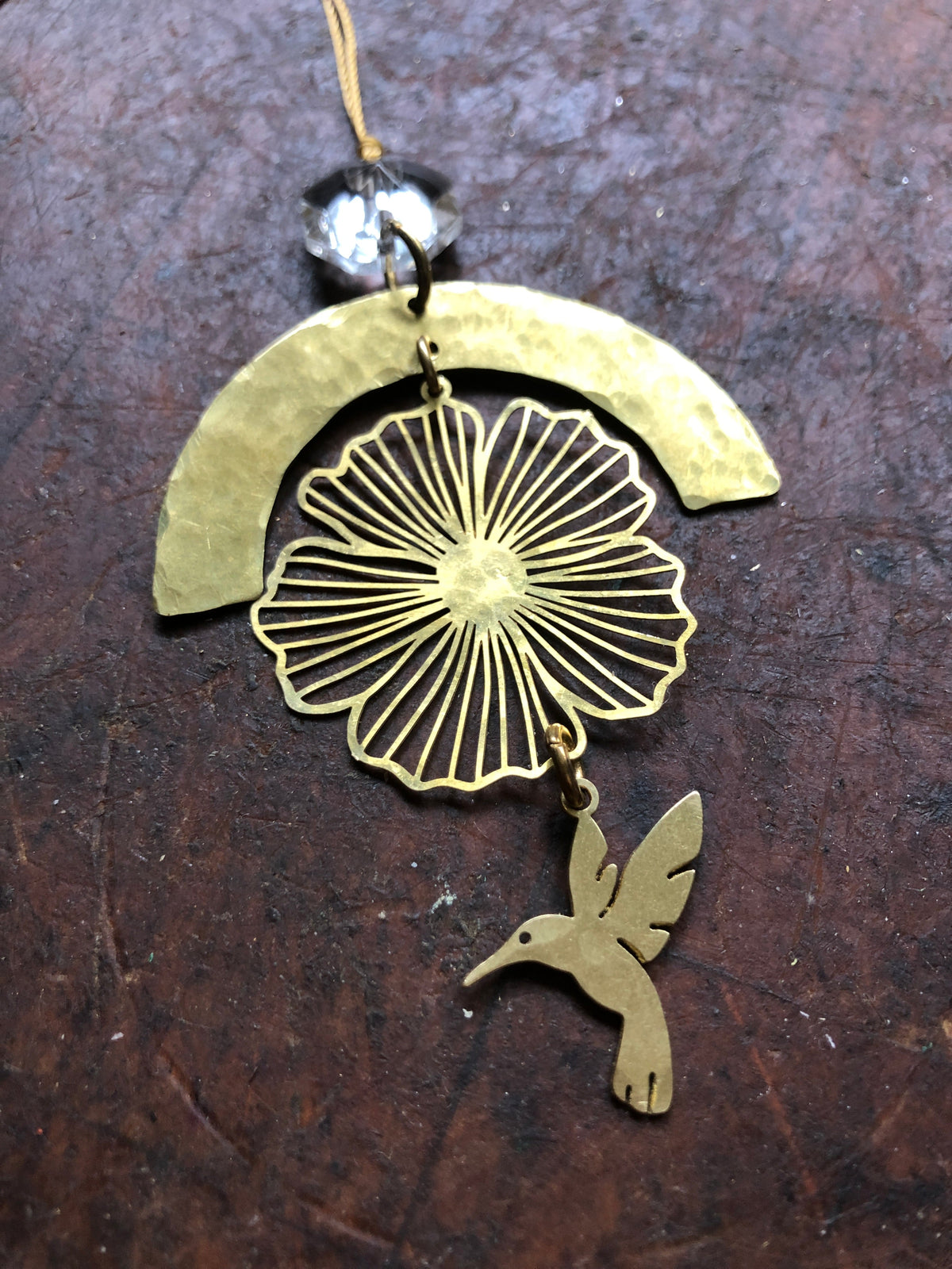 Brass ornament featuring a hammered semicircle, a floral wire design, and a small brass hummingbird charm, with a vintage glass crystal bead above it.