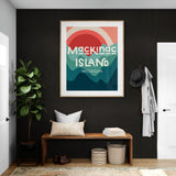 Framed modern art print with stylized sun and waves in red, coral, and teal hues, showing text 'Mackinac Island Michigan' on a dark wall above wooden bench.