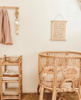 Round rattan baby bassinet with pale pink bedding in a minimalist beige nursery featuring a wooden shelf with folded linens and a wall hanging with script text.