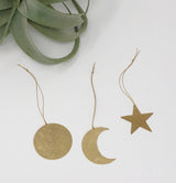 Three hammered brass ornaments shaped as a circle, crescent moon, and star, each with a hanging loop and string on a white surface beside green plant leaves.