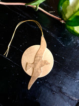 Smooth round wooden ornament with a bird silhouette cutout, shown on a dark textured surface with a green leafy plant nearby.