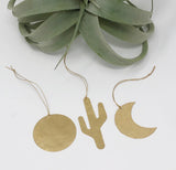 Three hammered brass ornaments shaped as a circle, cactus, and crescent moon, each with a thin string for hanging, displayed beneath a large green succulent leaf.