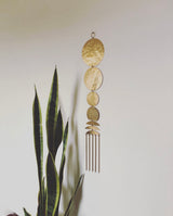 Brass-toned metal wall hanging with five connected hammered circular and crescent shapes, plus five vertical rods, hanging against a white wall beside snake plant leaves.