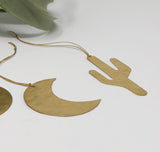 Two flat brass ornaments on a white surface, one shaped like a crescent moon and the other like a stylized cactus, both with textured surfaces and hanging cords.
