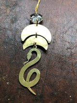 Serpentine brass ornament with a coiled snake design hanging below two semi-circular brass shapes, topped by a faceted vintage glass crystal against a textured dark surface.