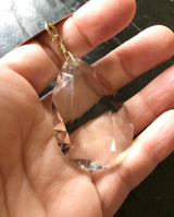 Hand holding a clear faceted crystal suncatcher pendant with a small gold wire loop attachment at the top against a dark background.