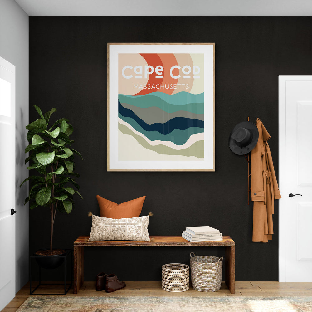 Abstract Cape Cod art print with layered waves in teal, navy, and beige under a minimalist sun in coral and cream, framed in light wood against a black wall.