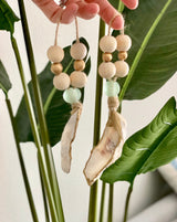 Two hanging sea-themed ornaments with wooden beads, light green glass beads, and oyster shell pieces, held against large green tropical leaves.