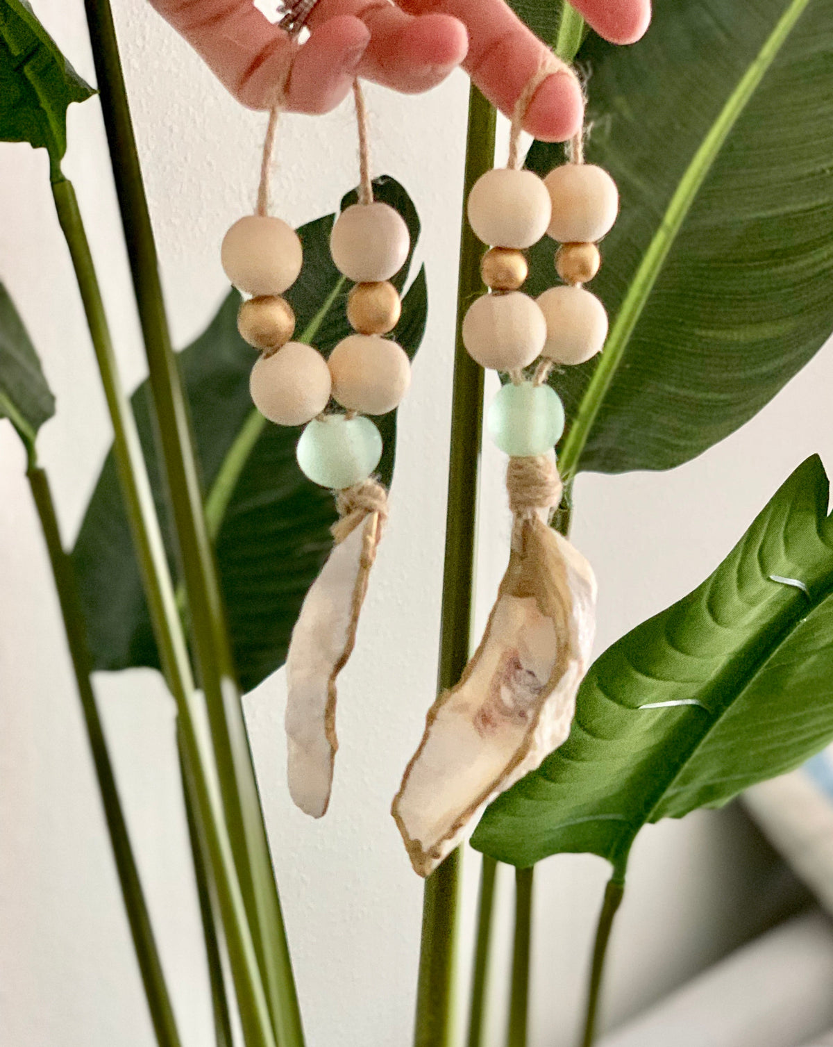 Two hanging sea-themed ornaments with wooden beads, light green glass beads, and oyster shell pieces, held against large green tropical leaves.