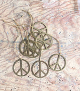 Seven brass peace symbol ornaments on natural twine strings, laid on a textured, light wood surface with saw marks and scattered black specks.