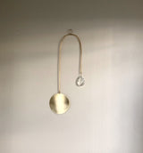 Minimalist suncatcher with a brushed gold circular disc and a gold curved wire ending in a clear faceted crystal prism, hanging against a plain light wall.