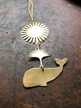 Gold-tone metal pendant featuring a stylized sunburst above a connecting fan-shaped element, with a whale silhouette charm at the bottom, all on a worn wooden surface.