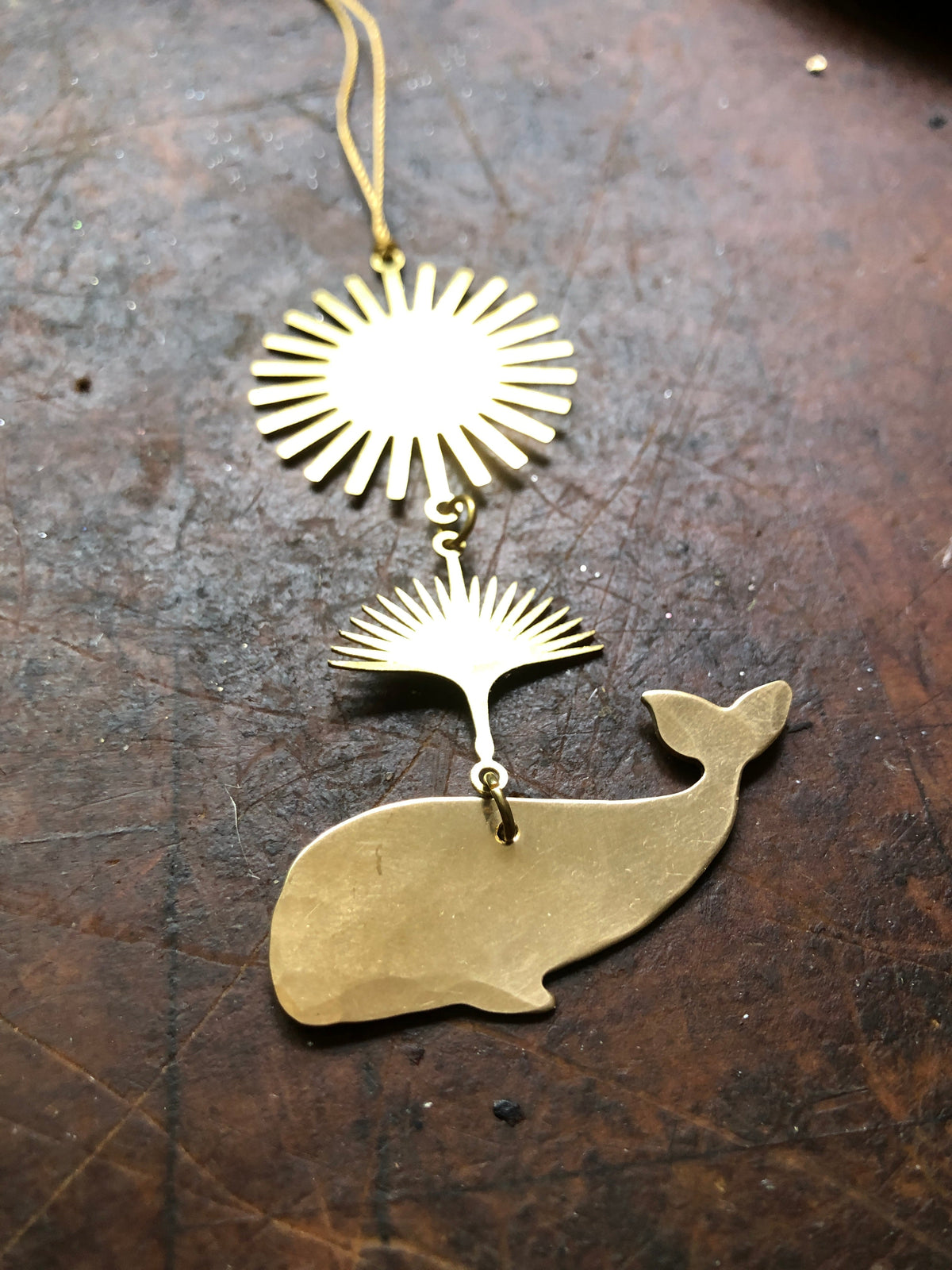 Gold-tone metal pendant featuring a stylized sunburst above a connecting fan-shaped element, with a whale silhouette charm at the bottom, all on a worn wooden surface.