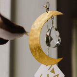 Gold-toned crescent moon metal piece with hammered texture holding a faceted clear crystal teardrop, suspended by twine with a geometric white paper charm below.