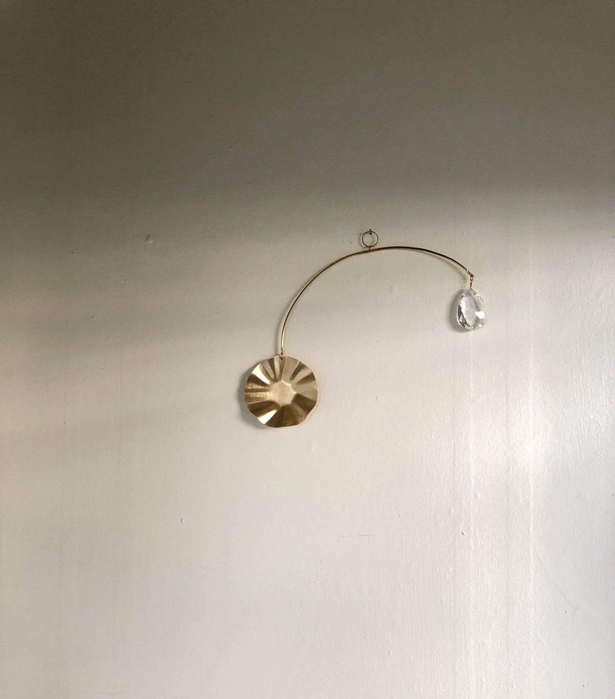 Minimalist wall suncatcher with a textured gold disc and a clear faceted crystal, suspended on a delicate curved gold wire against a white wall.