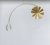 Minimalist brass mobile suncatcher with a circular hammered brass disc on one end and a faceted clear teardrop crystal on the other, suspended on a curved arm.