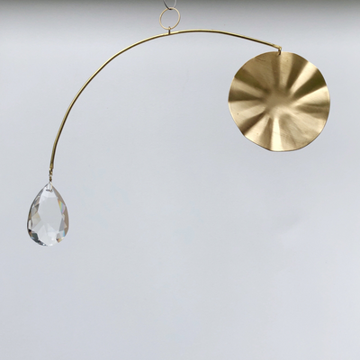 Aura Suncatcher made of hand-cut brushed brass and vintage glass crystal hanging indoors against a neutral wall