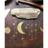 Wall sculpture with a rough driftwood piece at top and hanging hammered brass moon and stars in varying sizes, suspended by delicate gold chains on a brown background.