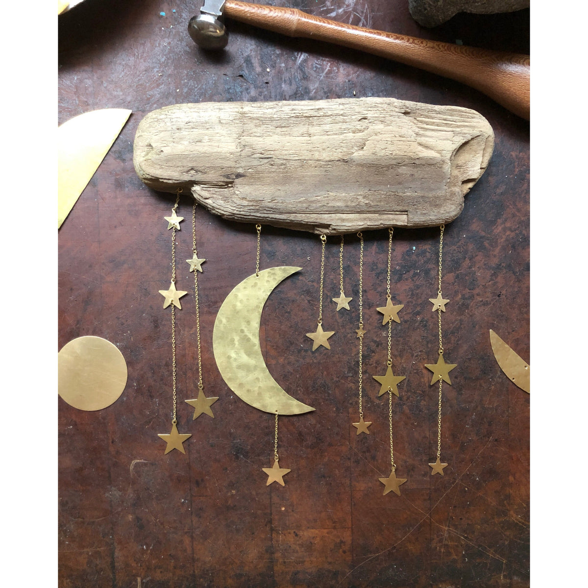 Wall sculpture with a rough driftwood piece at top and hanging hammered brass moon and stars in varying sizes, suspended by delicate gold chains on a brown background.