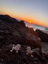 Pair of pearl earrings shaped like poodles resting on a dark rock at sunset with orange and blue sky hues and ocean waves in background.