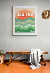 Framed modern art print with stylized layered waves in green and orange hues, labeled 'Lido Key Florida,' hanging above a wooden bench with a light gray throw.