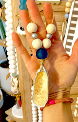 Hand holding a decorative hanging ornament with a large blue bead, white and silver beads, and a dangling shell with a pearly interior finish.