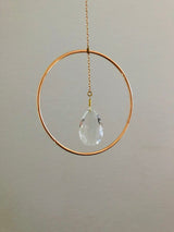 Teardrop-shaped clear crystal hanging inside a thin, gold circular hoop suspended by a delicate chain against a plain beige background.